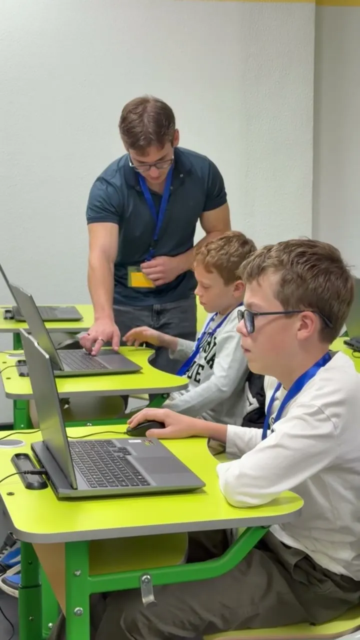 Profesorul le explică pașii de lucru unor elevi la laptopuri